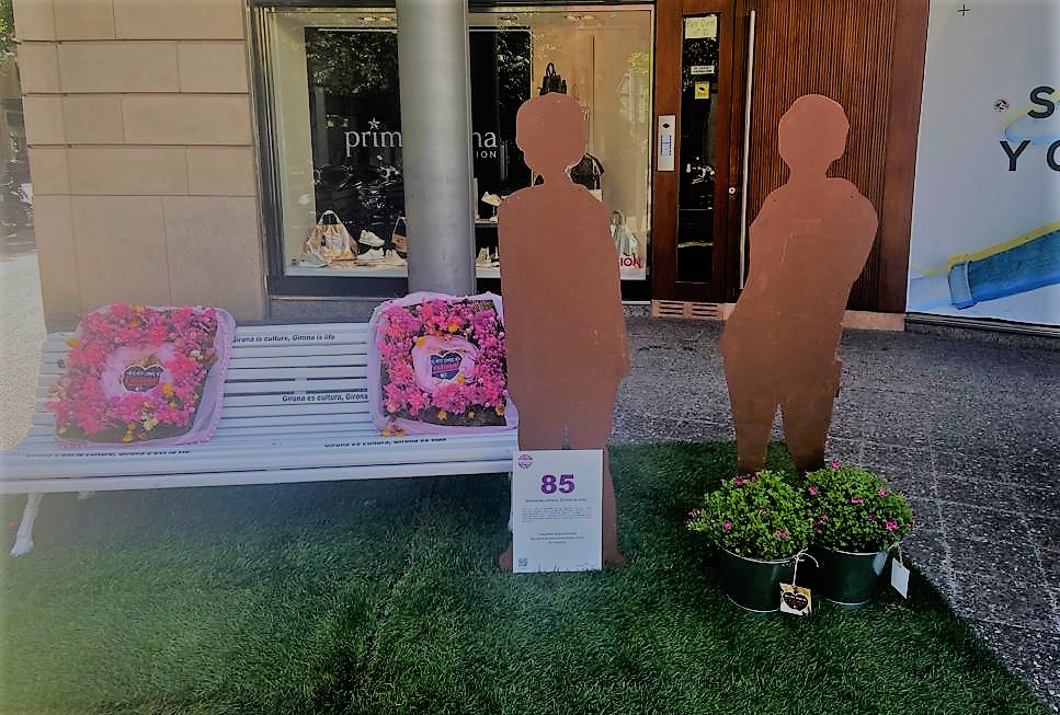 Un banc amb coixins fets amb flors i al costat dues siluetes de persones. Al darrere, tres persones
