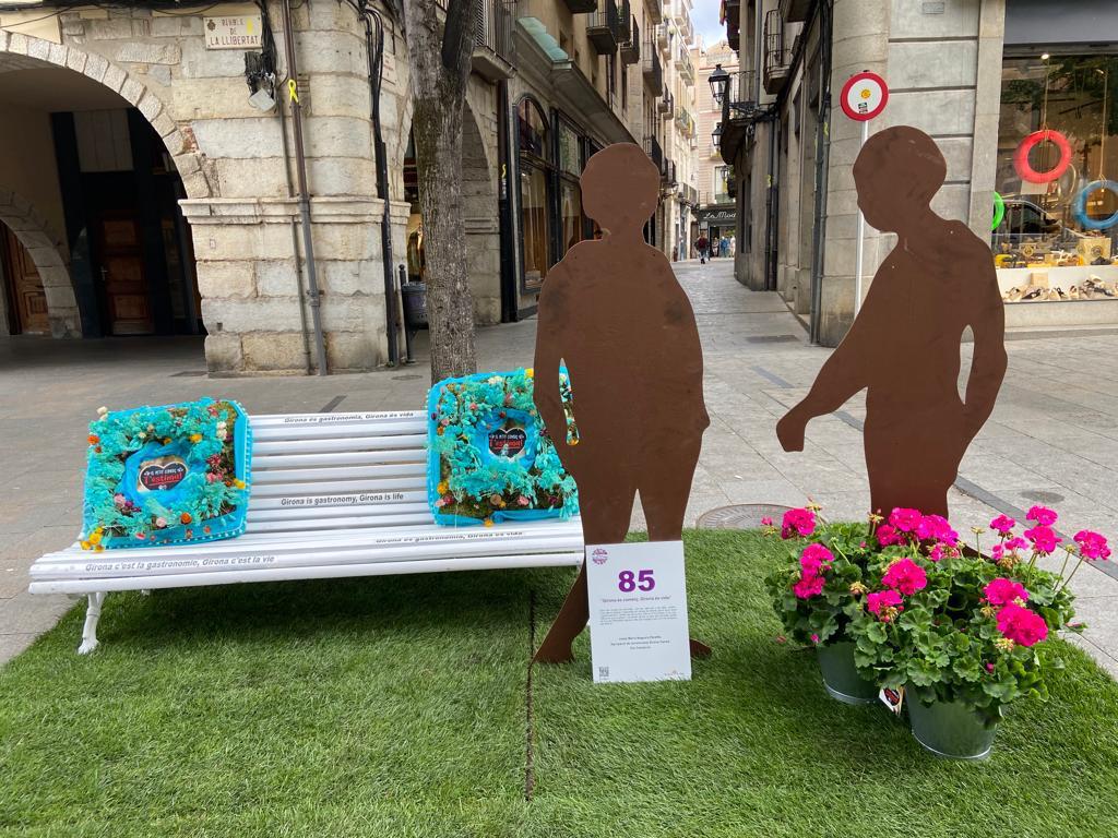 Un banc amb coixins fets de flors i al costat dues siluetes de persones sobre una catifa de gespa