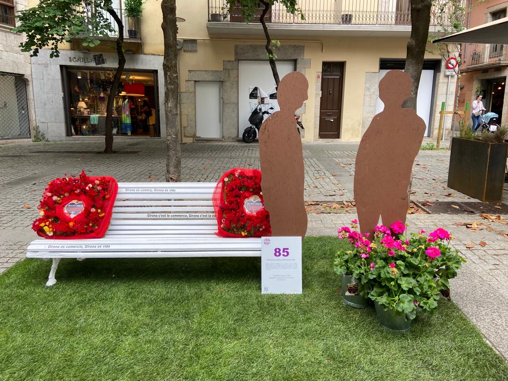 Banc amb coixins fets de flors i al costat dues siluetes de persones sobre una catifa de gespa