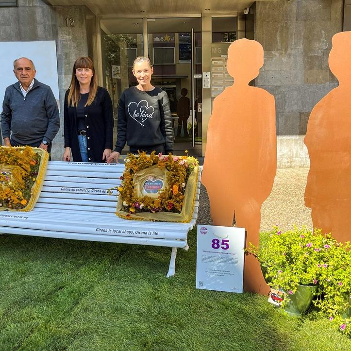 Un banc amb coixins fets amb flors i al costat dues siluetes de persones. Al darrere, tres persones