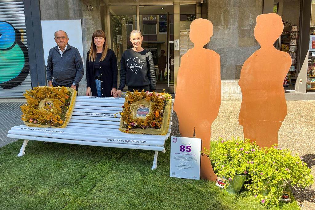 Un banc amb coixins fets amb flors i al costat dues siluetes de persones. Al darrere, tres persones