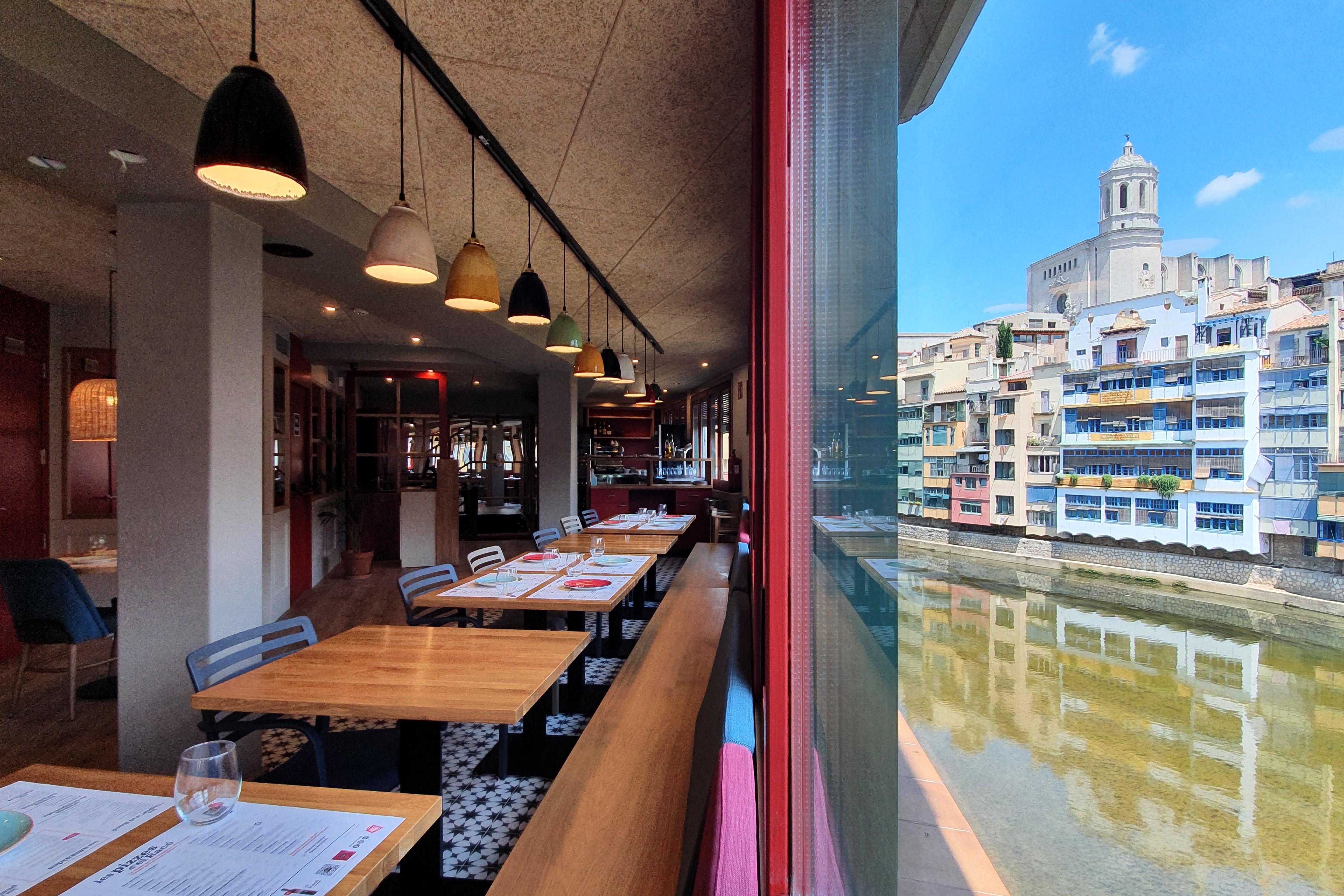 Visió del restaurant; a l'esquerra les taules, i la dreta una finestra a través de la qual es veu el riu, les cases  de colors, amb la catedral al fons