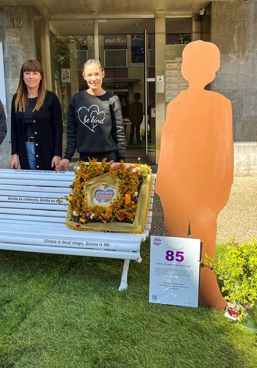 Un banc amb coixins fets amb flors i al costat dues siluetes de persones. Al darrere, tres persones